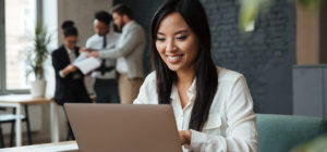 Picture of cheerful young asian business woman sitting indoors using laptop computer. Looking aside.